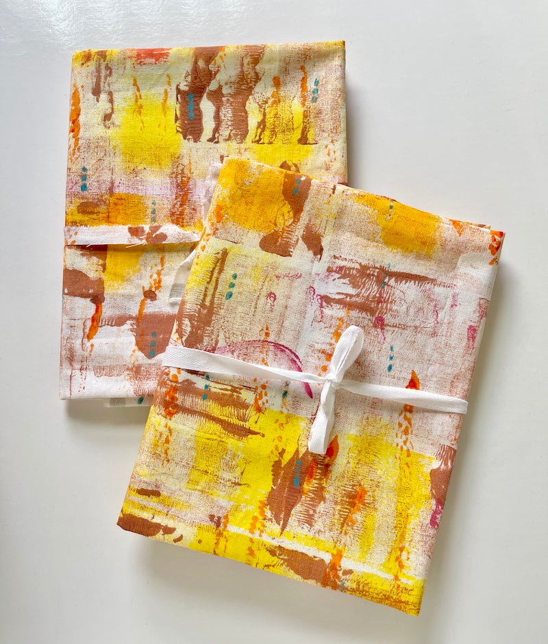 Two pieces of hand-painted fabric featuring yellow, orange, brown, pink  and blue paint arranged in an abstract design. Each piece of fabric is folded and tied with ribbon.