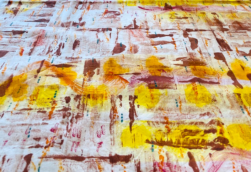 A large piece hand-painted fabric featuring yellow, orange, brown, pink  and blue paint in an abstract design . The fabric is laid out flat on a surface.